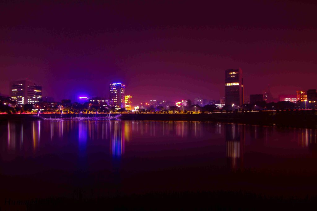 A cityscape at night with vibrant reflections of illuminated buildings on a calm water surface, showcasing a mix of purple, blue, and yellow hues.