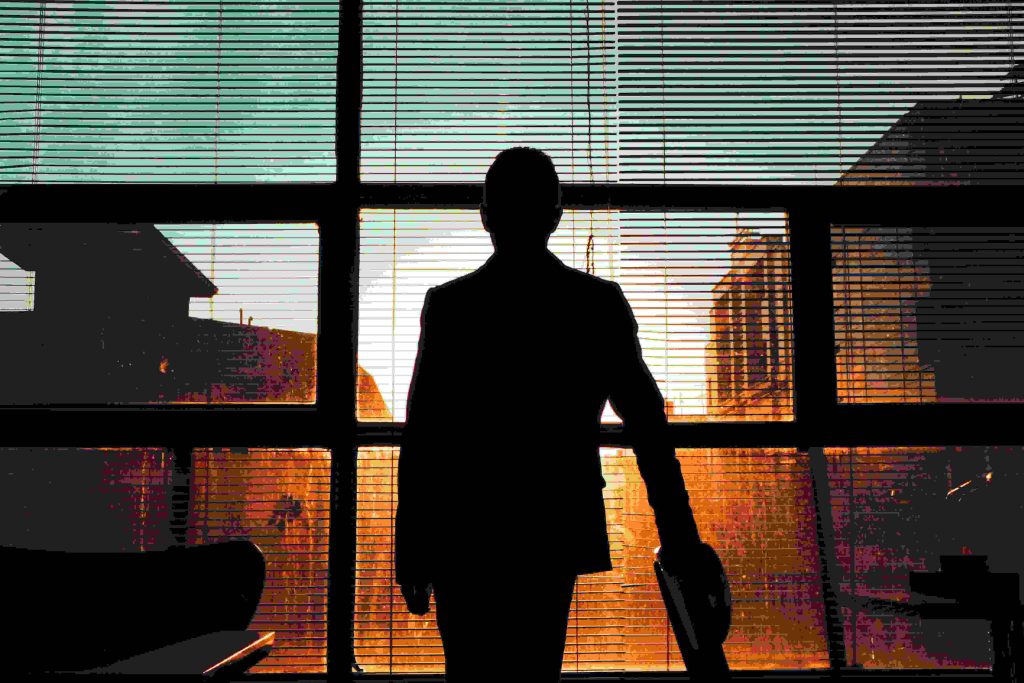  A silhouette of a person in formal attire standing by a large window with blinds, overlooking an urban scene bathed in a golden sunset