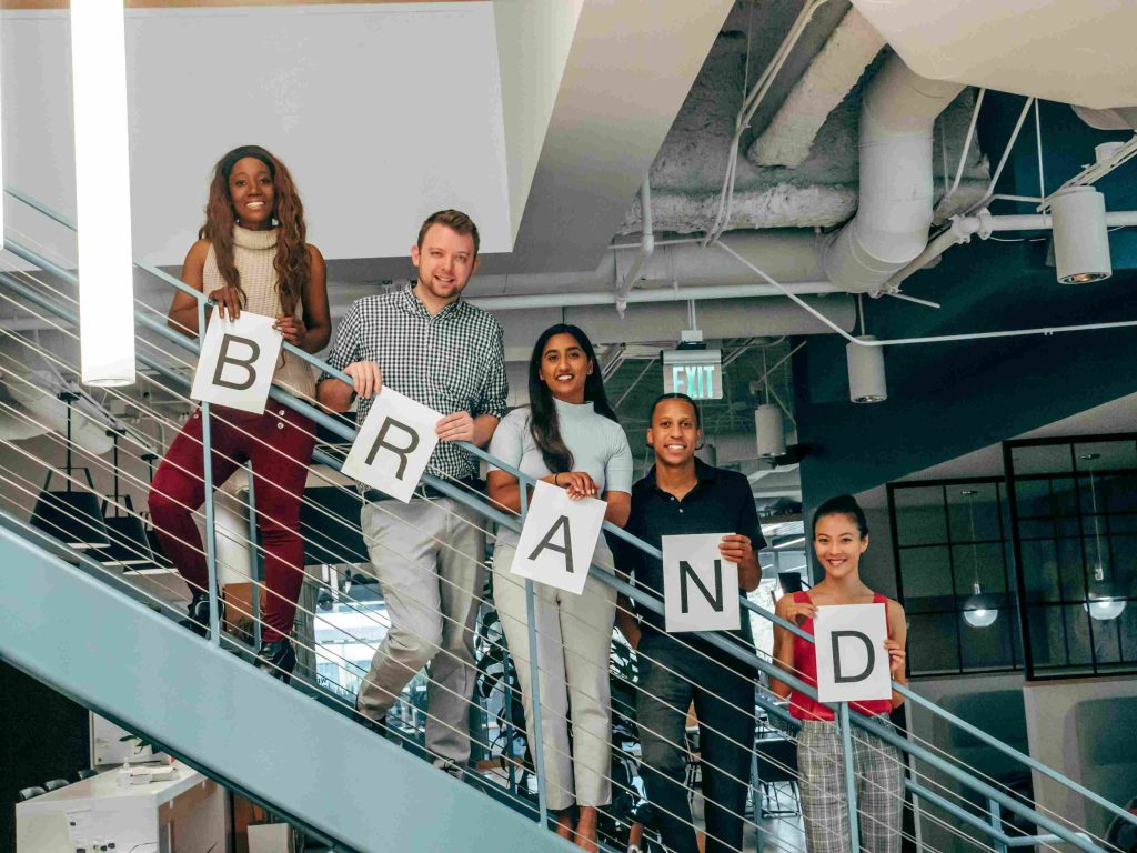 A diverse group of five professionals standing on a staircase in an office, each holding a letter to form the word 'BRAND.' The team is smiling and symbolizes collaboration, creativity, and teamwork in branding and marketing strategies.